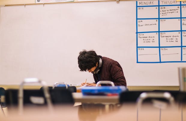 A young male adult sits writing at a desk in a classroom